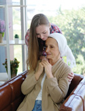 A younger woman gently embracing an older woman in a headscarf as they sit together on a sofa in a bright, peaceful room.