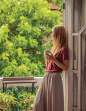 Woman standing by an open doorway holding a mug, looking out at lush green trees on a peaceful balcony.