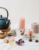 A collection of various crystals, lit pink candles, a singing bowl, and a teal teapot on a white surface.