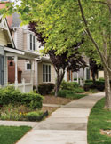 Quiet suburban neighborhood with tree-lined sidewalk, manicured lawns, and townhomes in soft daylight.