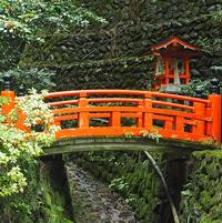Bright red wooden bridge with a small matching shrine, spanning a narrow moss-covered stream in a lush forest setting.
