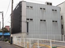 Current photo of where Hayashi Sensei's Reiki clinic was located in Toyko. Small gray three-story building with narrow windows and fenced parking area on a quiet street, utility pole and wires alongside.