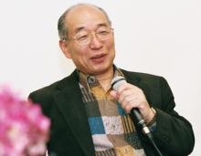 Hiroshi Doi Sensei wearing glasses and a patterned shirt is smiling and holding a microphone, seated against a plain background.