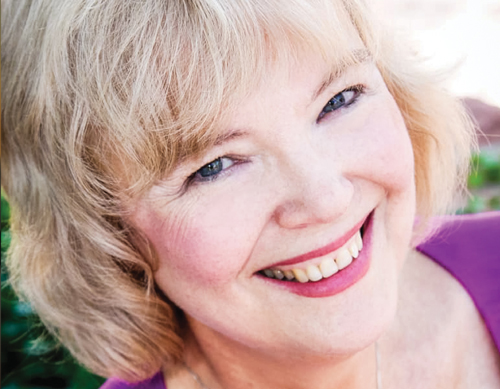 Close-up of a smiling woman with light blonde hair, outdoors in soft natural light.