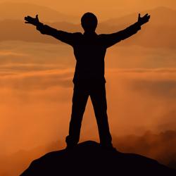 Silhouette of a person standing on a peak with arms outstretched against an orange sky and misty mountain background.
