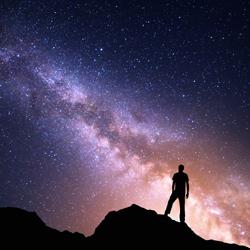 Silhouette of a person standing on a rocky ridge at night, gazing at the bright, colorful Milky Way stretching across a star-filled sky.