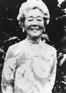 Hawayo Takata Black-and-white photo of Hawayo Takata smiling, wearing a patterned dress, standing outdoors in front of dark foliage.