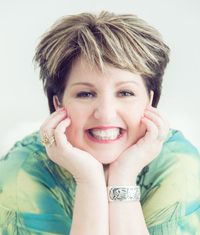 Smiling woman with short brown and blonde hair, hands on cheeks, wearing a green top and silver bracelet.